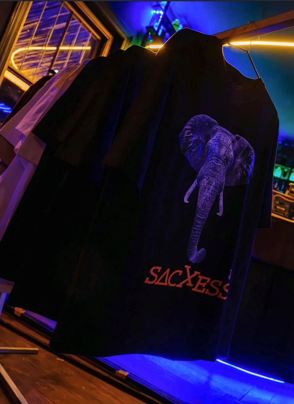 The SACXESS elephant graphic on a black tee, hanging on a rail under neon light